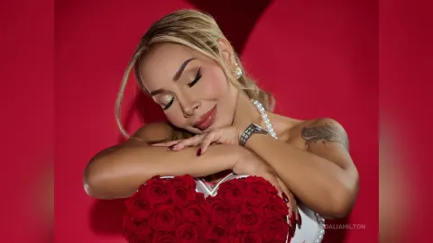 DaliaHilton — DaliaHilton smiles with closed eyes while hugging a large heart-shaped bouquet of red roses against a red background.