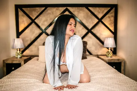 Smiling woman in a white shirt sitting on a bed in a cozy, warmly lit bedroom.