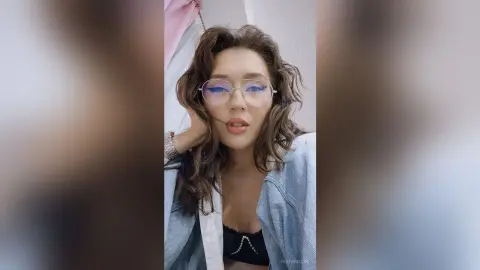 Young woman with wavy hair and glasses wearing a denim jacket in a soft, natural indoor setting.