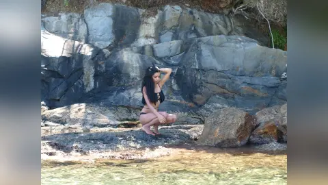 Young woman in black bikini sitting on rocky shore by clear ocean water under bright sunlight.