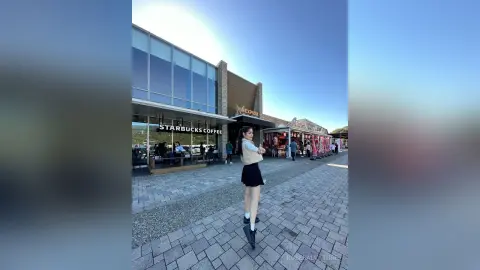 Young woman in a white blouse and black skirt walking past a Starbucks storefront on a sunny urban sidewalk.