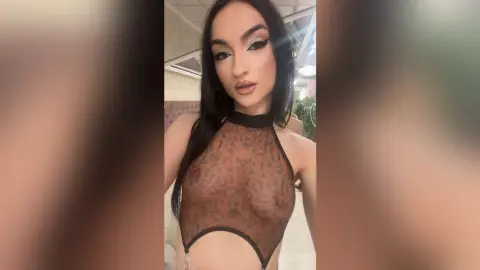 Glamorous woman in a sheer brown patterned halter top posing with dramatic makeup in a modern indoor setting.