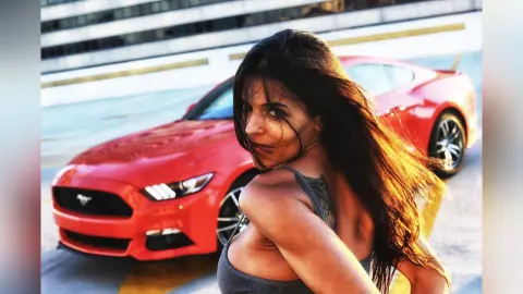 DrellaWolf — Stylish woman in a grey tank top posing confidently in front of a red Ford Mustang in an urban setting.