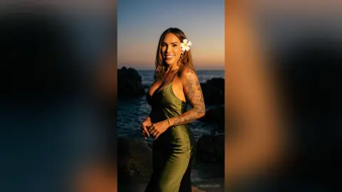 LonisMillers smiles while wearing a tight green satin dress and a white flower in her hair, posing outdoors by the ocean at sunset.