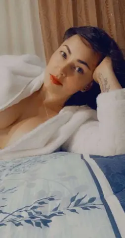 Woman in a white fluffy bathrobe relaxing on a patterned bed in a cozy, serene bedroom setting.