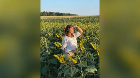 VictoriaWynn stands in a field of sunflowers, wearing a white button-down shirt and tan shorts, shielding her eyes from the sun.