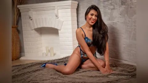 SamanthaHarvey sits on a grey shag rug in front of a white fireplace wearing a blue lace bodysuit.