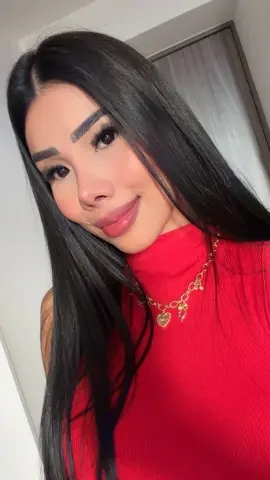 Smiling young woman with long dark hair wearing a red turtleneck and gold heart necklace in a bright room.