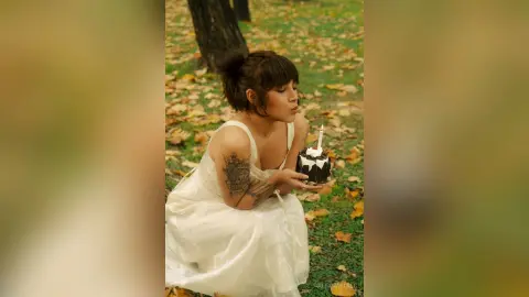 AlexiaVitali sits in a park with autumn leaves, wearing a white dress and holding a small chocolate cake with a single lit candle.