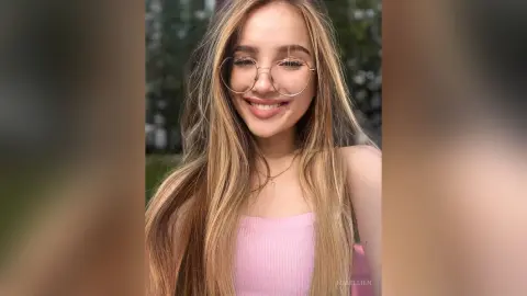 Smiling young woman with long blonde hair and round glasses wearing a pink ribbed top in soft natural sunlight.