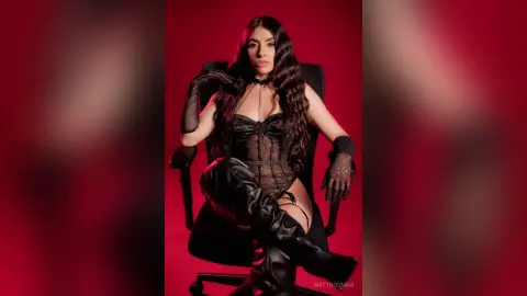 Glamorous woman in black corset and leather boots sitting on a chair against a red background.