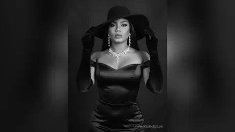 Elegant woman in a black satin dress and wide-brimmed hat posing glamorously in a moody black and white portrait.