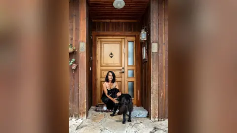 MichaelaBelle sits on a step in a black tank top and black shorts, posing next to a black dog in a wooden doorway.