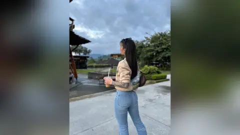 Woman in casual beige top and blue jeans holding a drink at an outdoor cafe under a cloudy sky.
