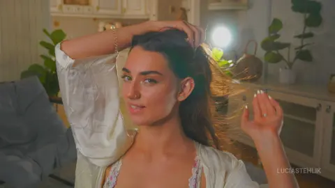 Young woman in white silk pajamas smiling in a cozy, sunlit bedroom setting.