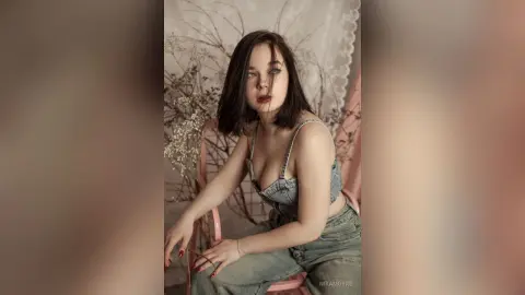Young woman in denim overalls sitting in a dreamy, floral indoor setting with a moody aesthetic.