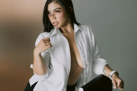 Young woman with glasses in an unbuttoned white shirt posing in a minimalist studio setting.