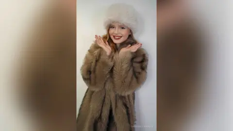 Smiling young woman in a luxurious brown faux fur coat and white fluffy hat posing against a white background.
