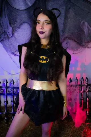 Woman in a Batgirl cosplay costume with gold accents posing in a spooky, dark Halloween setting.