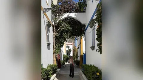 Woman in a white tank top walking through a sunny, narrow Mediterranean alleyway with white and yellow buildings.