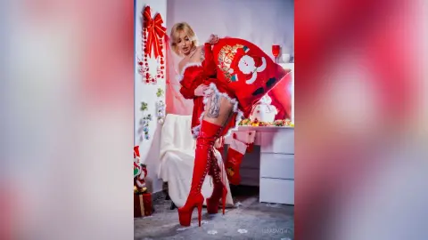 Blonde woman in a festive red Santa outfit and tall boots posing playfully in a decorated Christmas setting.