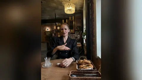 Smiling woman in a black blazer enjoying breakfast pastries at a cozy cafe table.