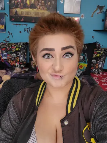 ChloeNightmare pouts her lips with a lip piercing, wearing a dark jacket and showing cleavage against a blue wall with horror-themed decor.