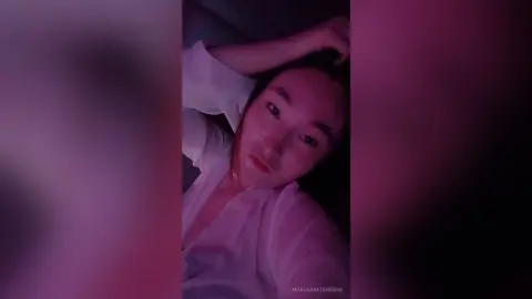 MaragaretSvedine lies on a dark surface wearing a white shirt under dim pink lighting.