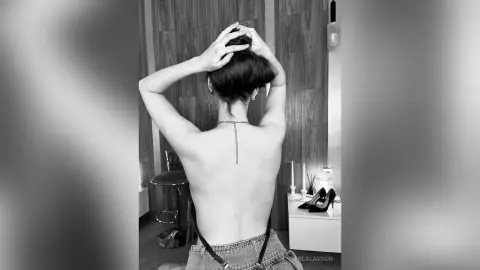 Back view of a woman in a black strappy top and denim jeans posing in a modern bedroom setting.