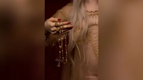 EllieFrosty holds a gold rosary against her chest, wearing a sheer, gold-trimmed lace bodysuit or slip.