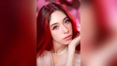 Young woman with vibrant red hair and winged eyeliner wearing a white pearl necklace in a soft pink setting.