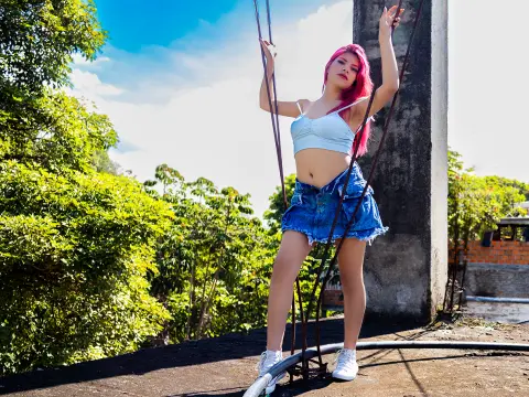 Young woman with pink hair in a light blue crop top and denim shorts posing on a swing outdoors in sunlight.