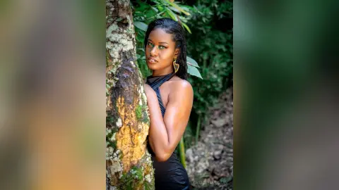 Elegant woman in a black sleeveless top leaning against a mossy tree in a lush green forest setting.
