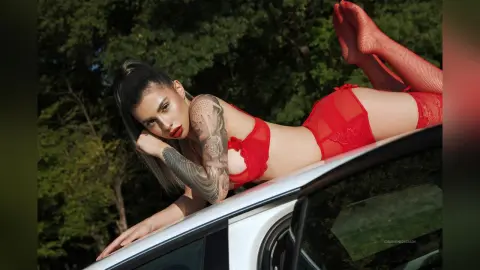 Woman in red lace lingerie relaxing on a white car roof against a lush green forest background.