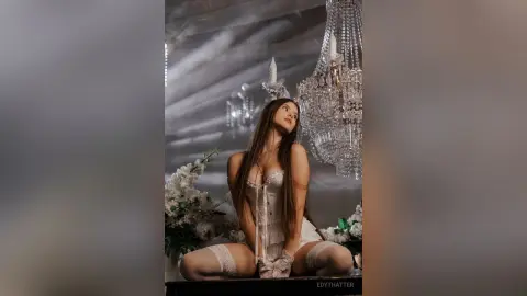 Elegant woman in white lace lingerie sitting in a dreamy, moody room with crystal chandeliers and white flowers.