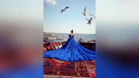 Elegant woman in a flowing blue sequin gown sitting on a rug overlooking a coastal city with birds flying above.