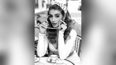 Elegant woman in white dress and polka dot headband sitting at a cafe table in a vintage black and white portrait.