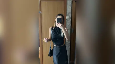 Young woman in a chic black sleeveless top and skirt posing for a mirror selfie in a modern hallway.