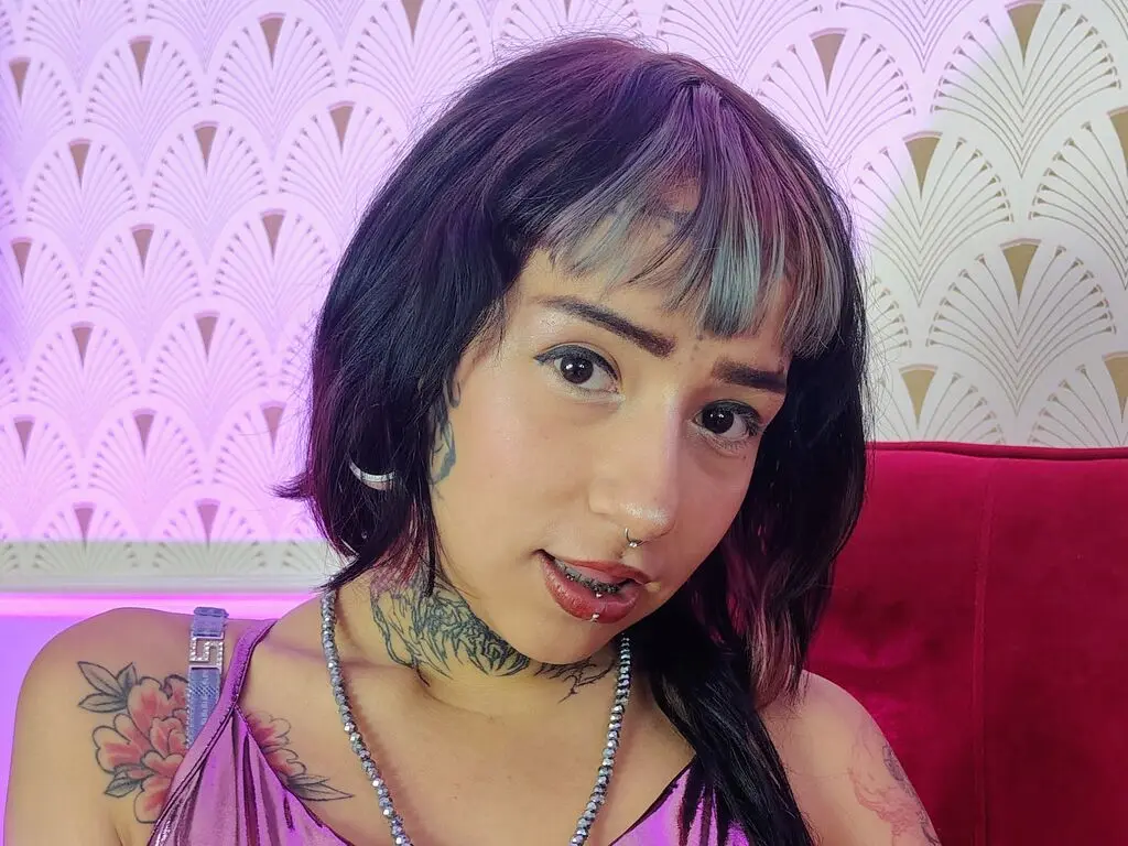 Young woman with dark bangs and tattoos wearing a pink top in a room with patterned wallpaper.