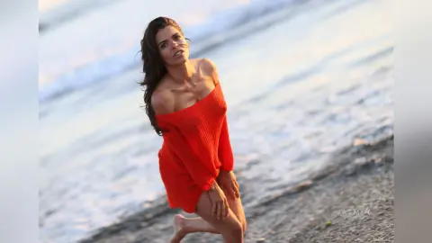 Beautiful woman in an orange knit dress posing on a sunny beach by the ocean waves.