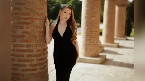 RubySoul poses outdoors in a black low-cut dress, leaning against a large brick column with her long wavy hair down.