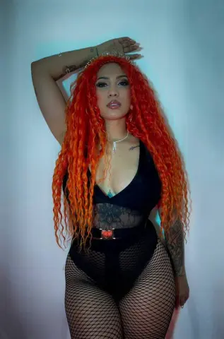 Woman with long wavy orange hair in a black bodysuit and fishnet tights posing against a white background.