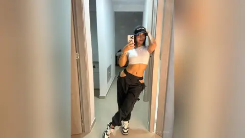Stylish woman in white crop top and black cargo pants taking a mirror selfie in a modern minimalist apartment.
