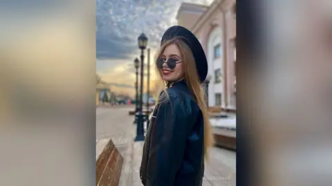 Smiling blonde woman in black leather jacket and sunglasses walking through a city street at sunset.