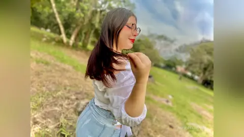 Young woman in a white blouse and denim shorts smiling in a lush green park under a cloudy sky.