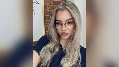 Blonde woman with glasses and navy blue shirt posing in a modern room with brick walls.