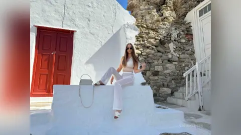 Stylish woman in white summer outfit sitting by a rustic stone wall and red door in a sunny Mediterranean village.