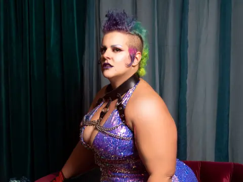Woman with colorful punk hair in a sequined purple outfit and leather choker against teal curtains.