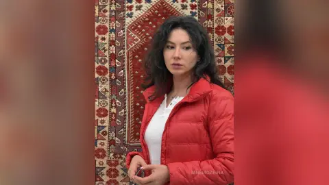 MnemoZeina has long dark hair and wears a red puffer jacket over a white shirt, standing in front of a patterned red rug.