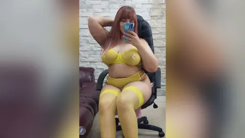 AdelineDevin poses in a black chair wearing a yellow bra, yellow panties, and matching yellow thigh-high stockings against a white brick wall.